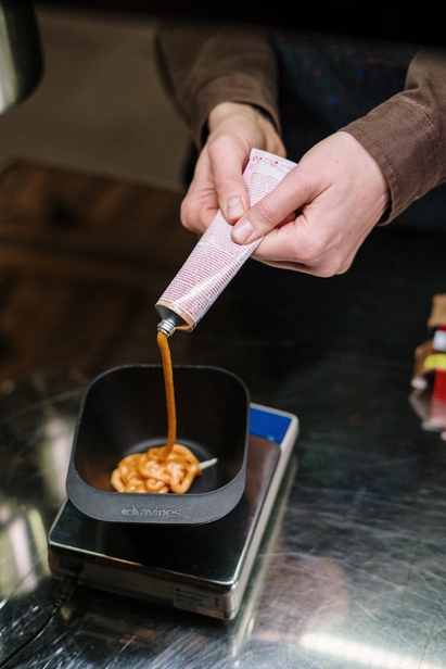 Hands squeezing tube of hair dye into a black mixing bowl on a digital scale